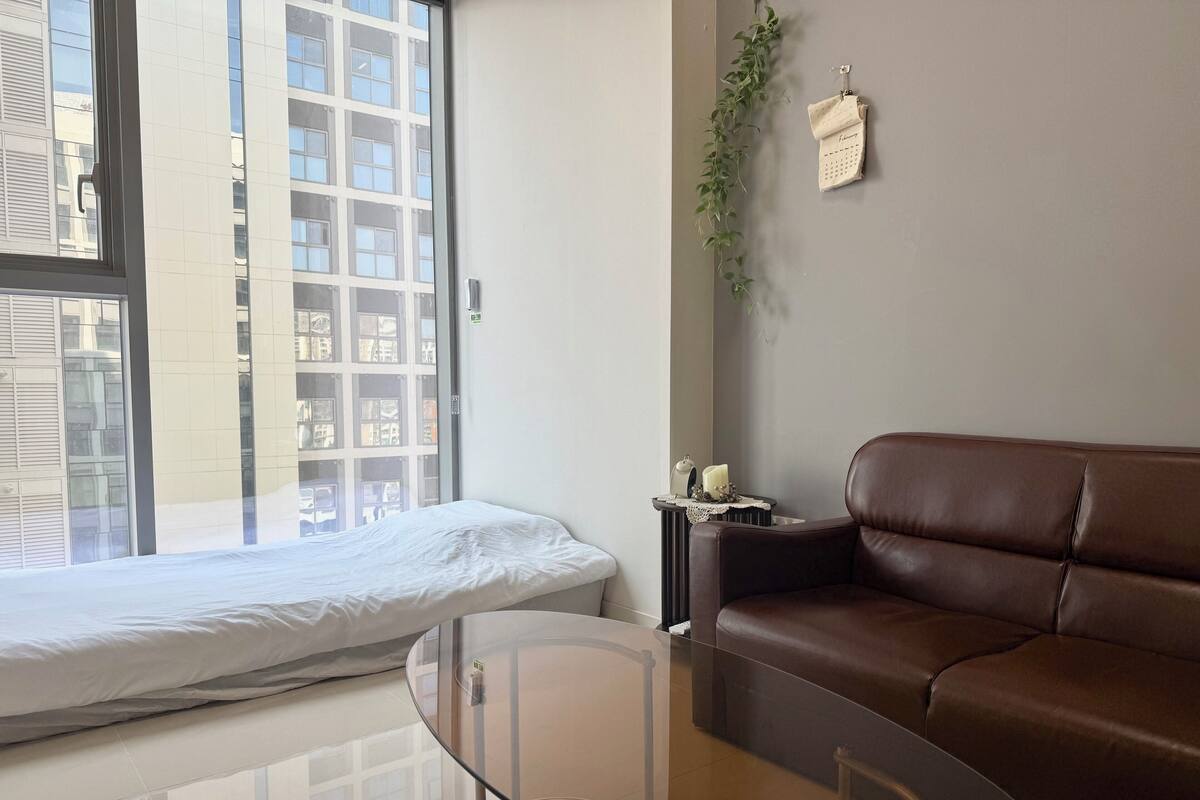 A modern living room with a glass coffee table, a brown leather sofa, a small side table with a candle, and a view of a building through a large window.