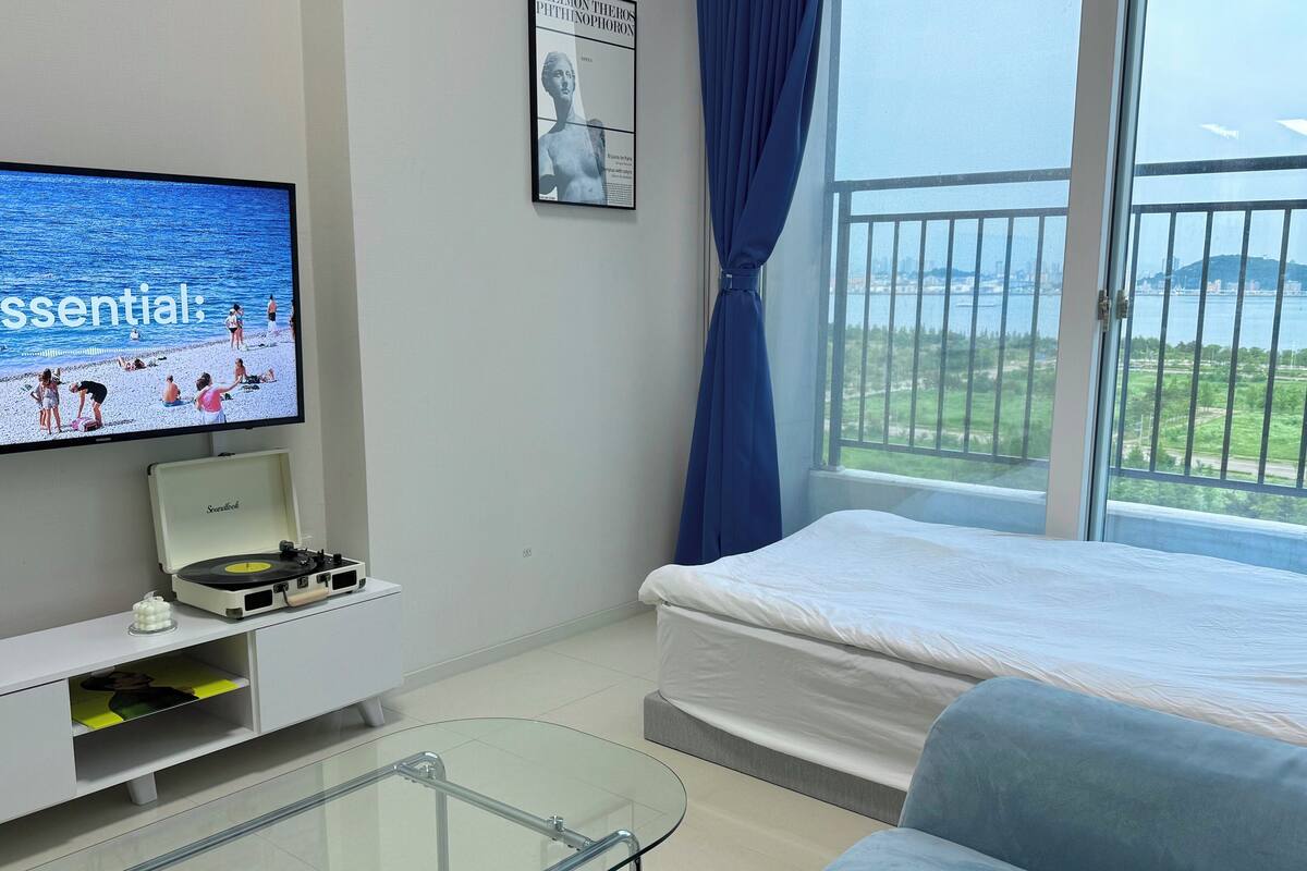 A modern hotel room with a bed, a glass coffee table, a television displaying a beach scene, a small music player, and a view of the sea through a large window.