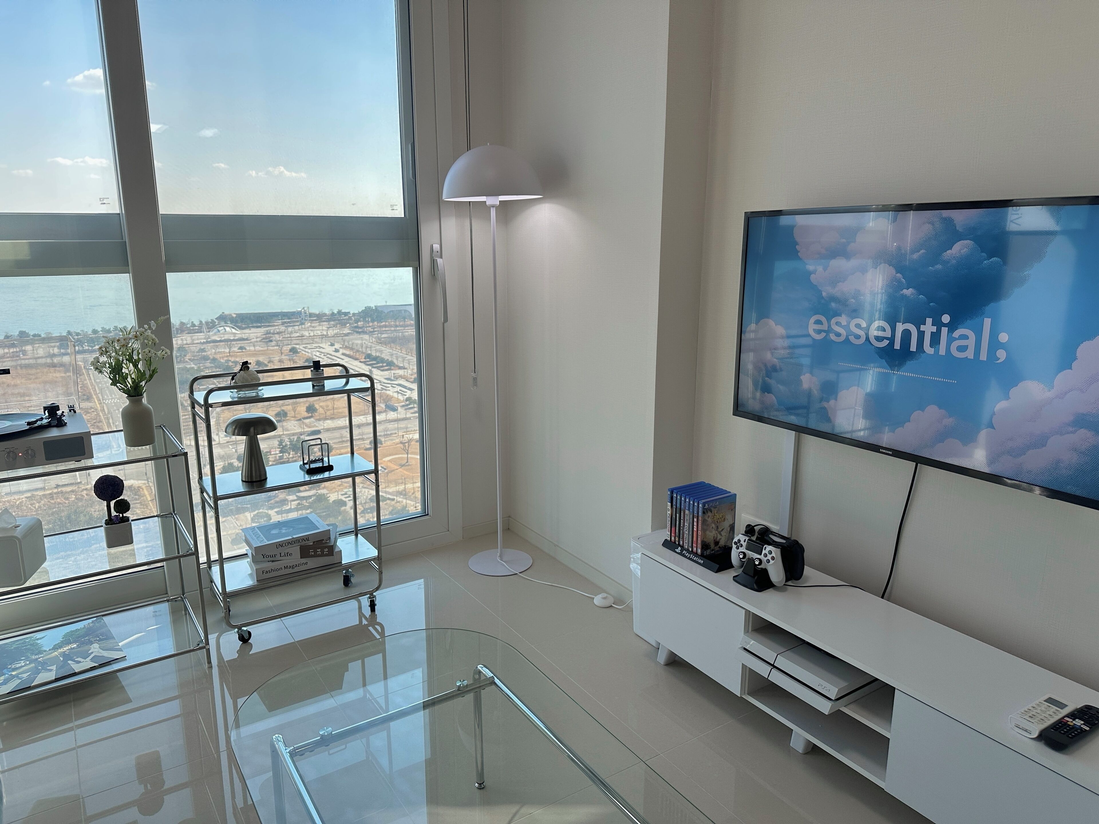 A modern living room with a large TV, a glass coffee table, a white console table, and a view of a cityscape through large windows.