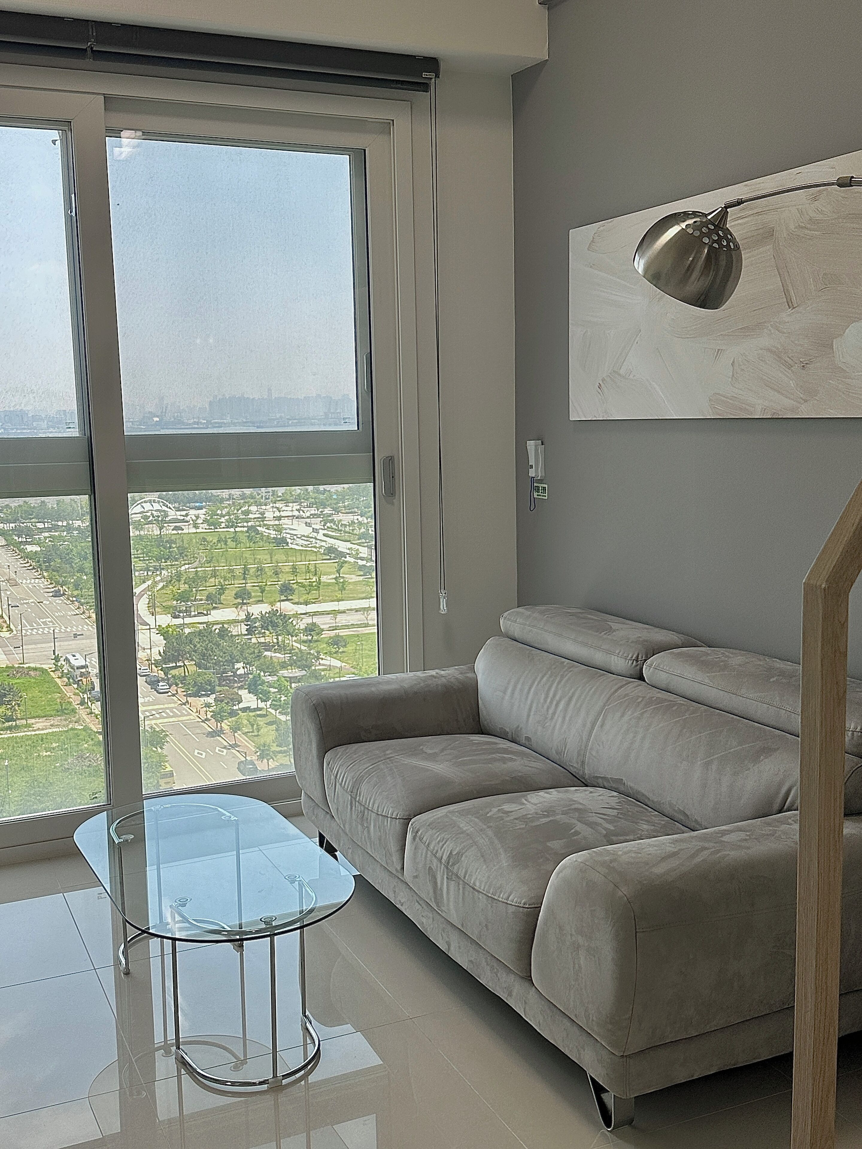 A modern living room with a grey sofa, a glass coffee table, and a large window offering a city view.