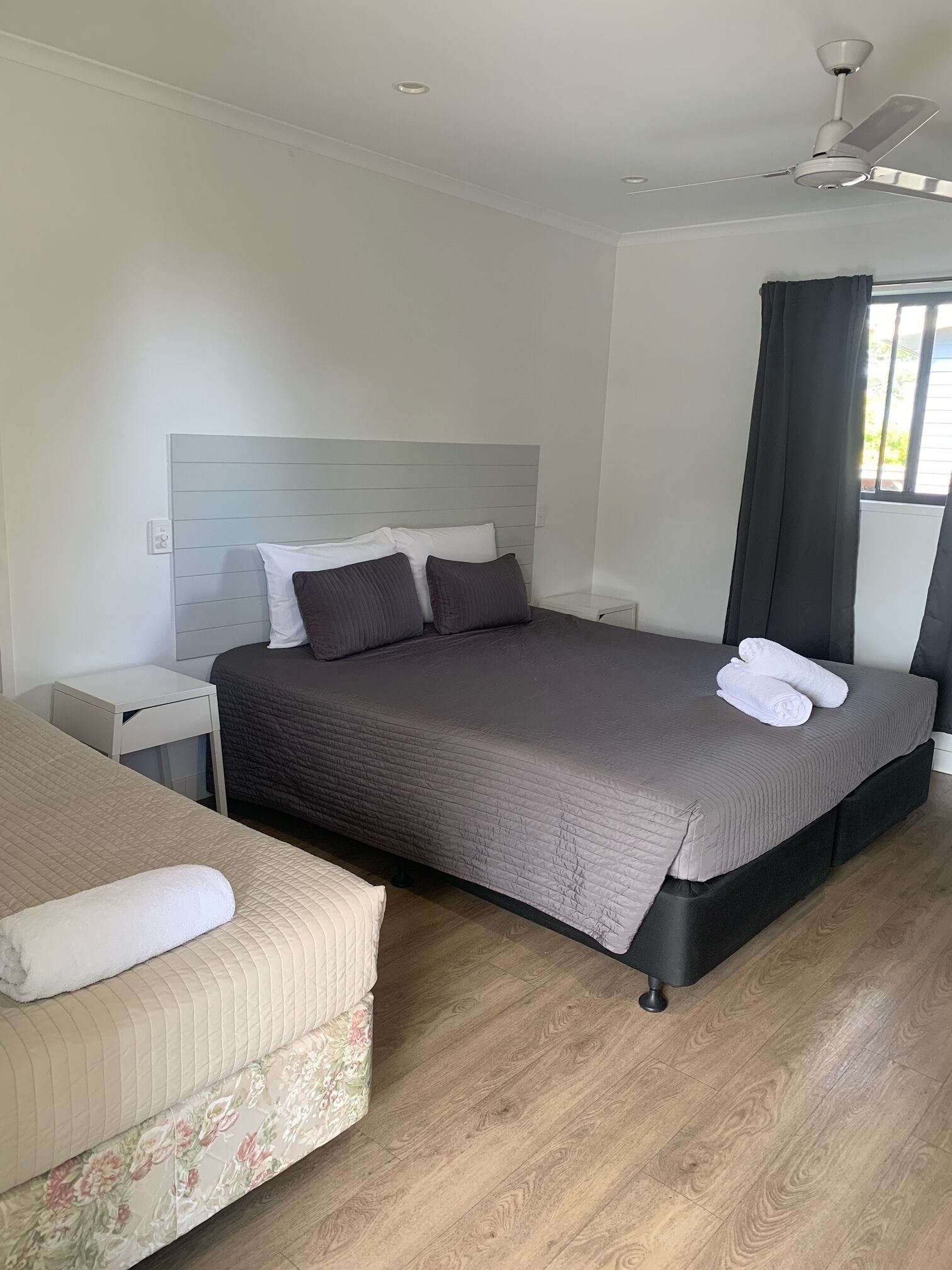 Ein modernes Schlafzimmer mit einem großen Bett, einer kleinen Sitzecke, einem Nachttisch, einem Deckenventilator und einem Fenster mit Vorhängen.