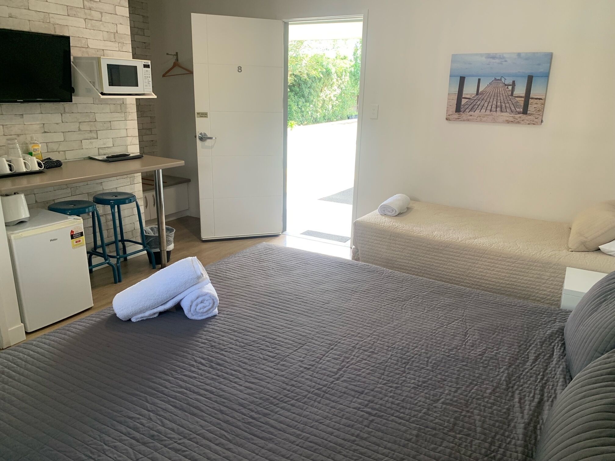 A hotel room with a bed, a small kitchenette, a TV, and a painting of a beach.