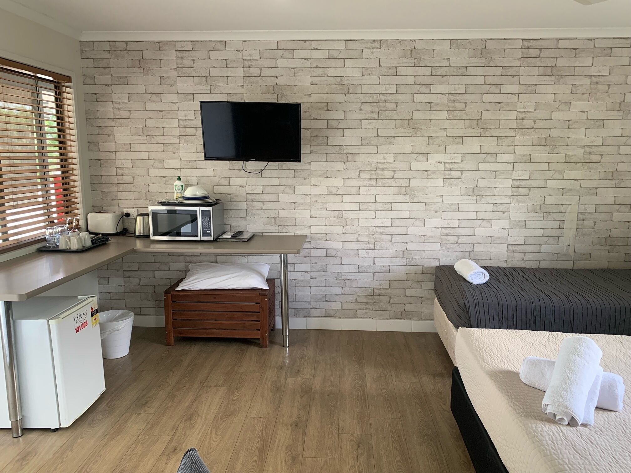 Una habitación de hotel compacta con pared de ladrillo, un televisor de pantalla plana sobre una kitchenette y una cama con colcha blanca, junto con un pequeño taburete de madera.