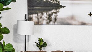 A living room with a brown sofa, a lamp, a small plant, and a framed picture of a lake with trees and mist.