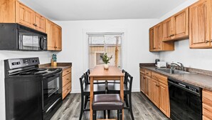 A kitchen with wooden cabinets, a black stove, a dining table with chairs, and a window with blinds.