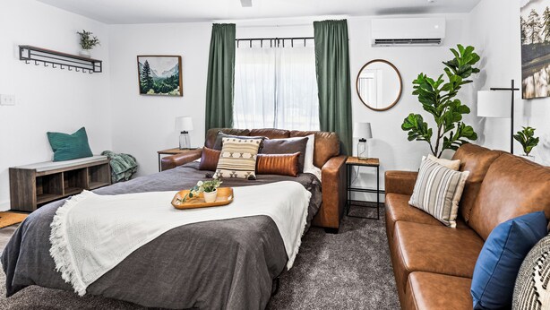 A modern bedroom with a bed, a brown leather sofa, a small side table, a bedside table, a ceiling fan, an air conditioning unit, a window with curtains, and a wall-mounted shelf with a plant.