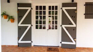 A white double door with a geometric design, flanked by two potted plants, and a doormat with a floral design. - Enjoy the peace and quiet of nature in this fresh, bright villa. (Saint-Joseph)
