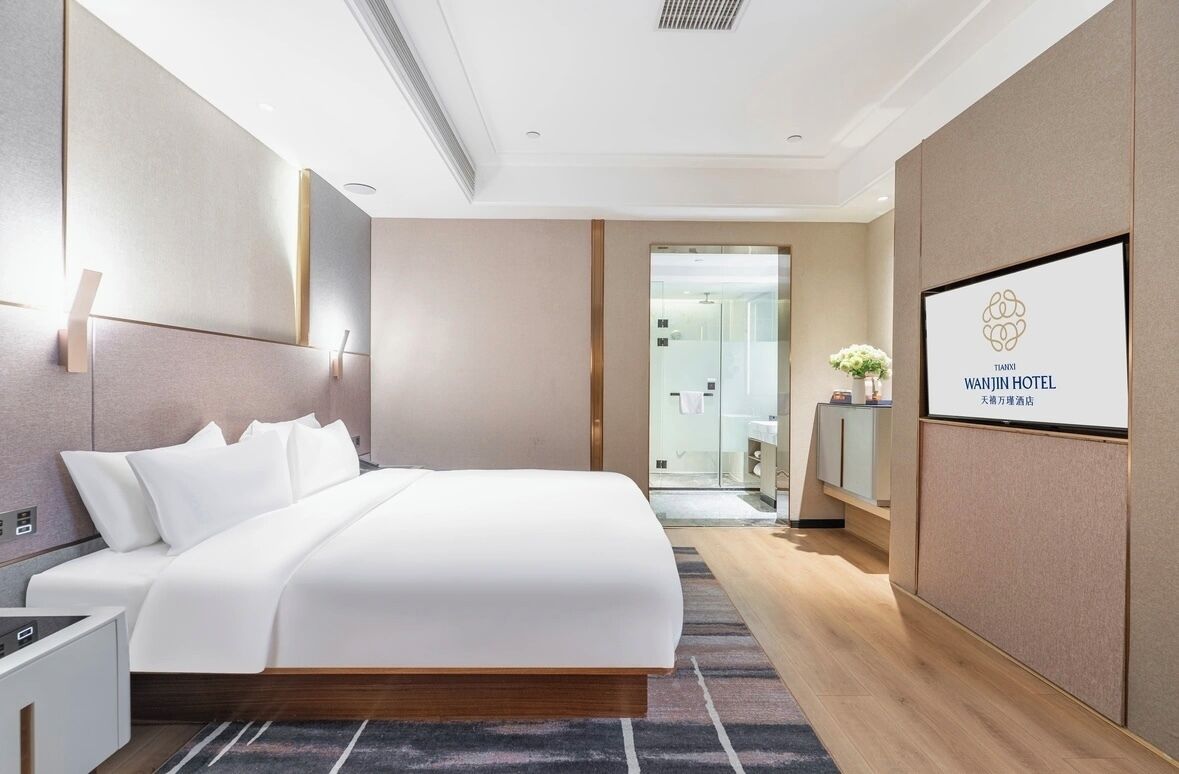 A modern hotel room with a large bed, a bedside table, a nightstand, a mirror, and a framed logo on the wall.