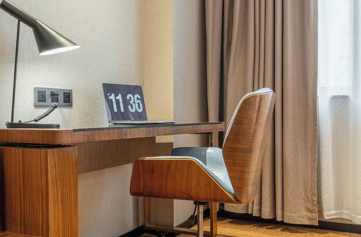 Et hotelværelse med et træskrivebord, en stol, en skrivebordslampe, et skilt med værelsesnummer 1136 og et vindue med gardiner.