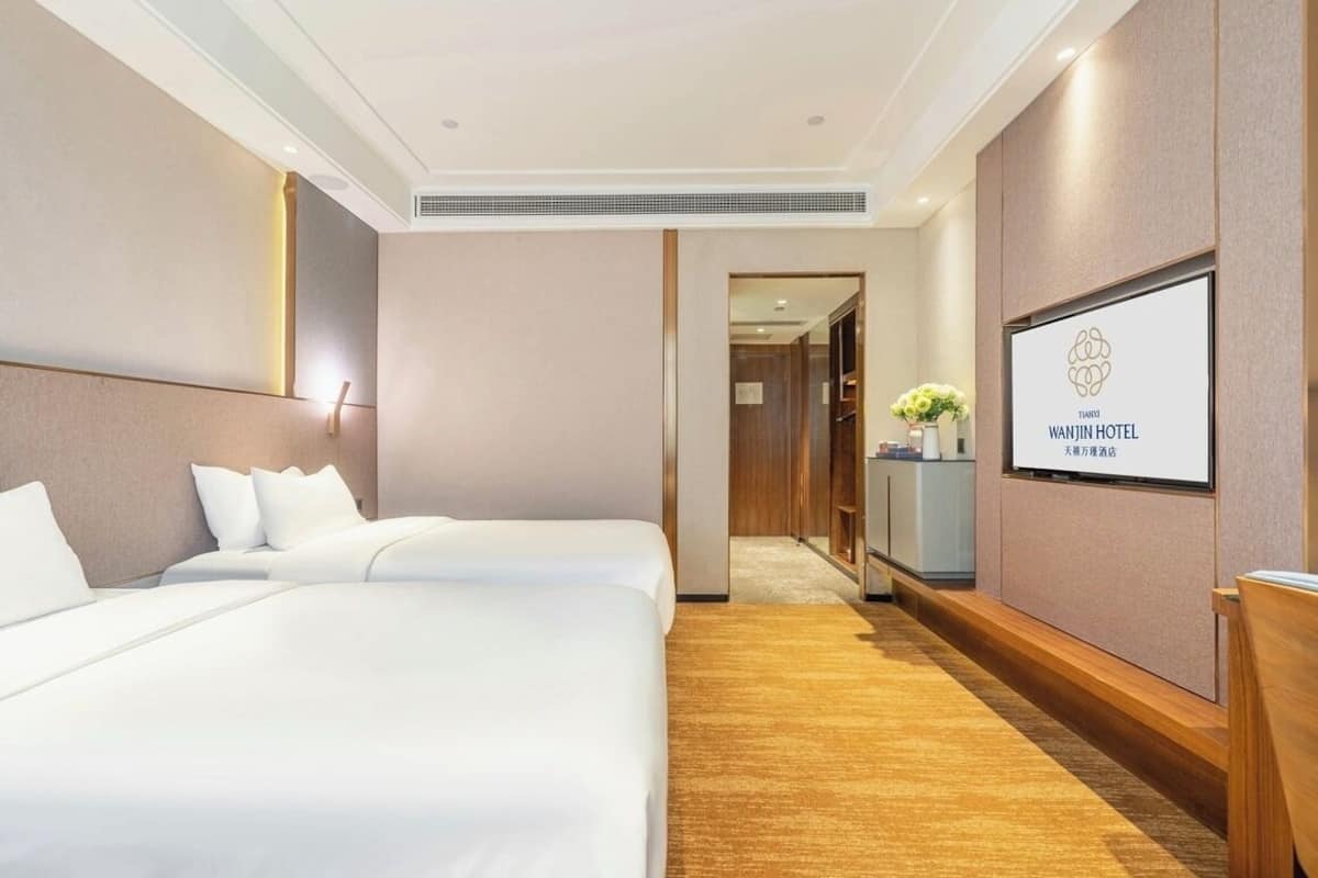 A hotel room with two beds, a wooden floor, a framed sign on the wall, and a hallway leading to another room.
