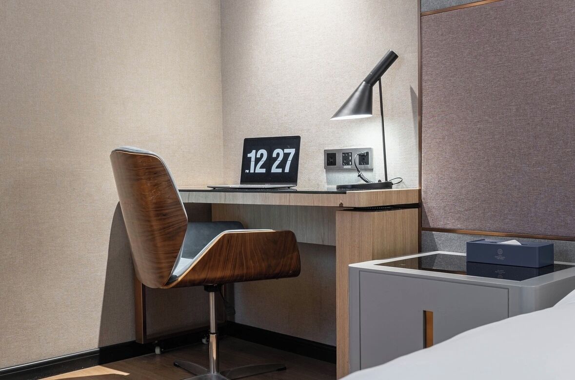 A modern hotel room with a wooden desk, a chair, a desk lamp, a clock, and a bedside table with a box.