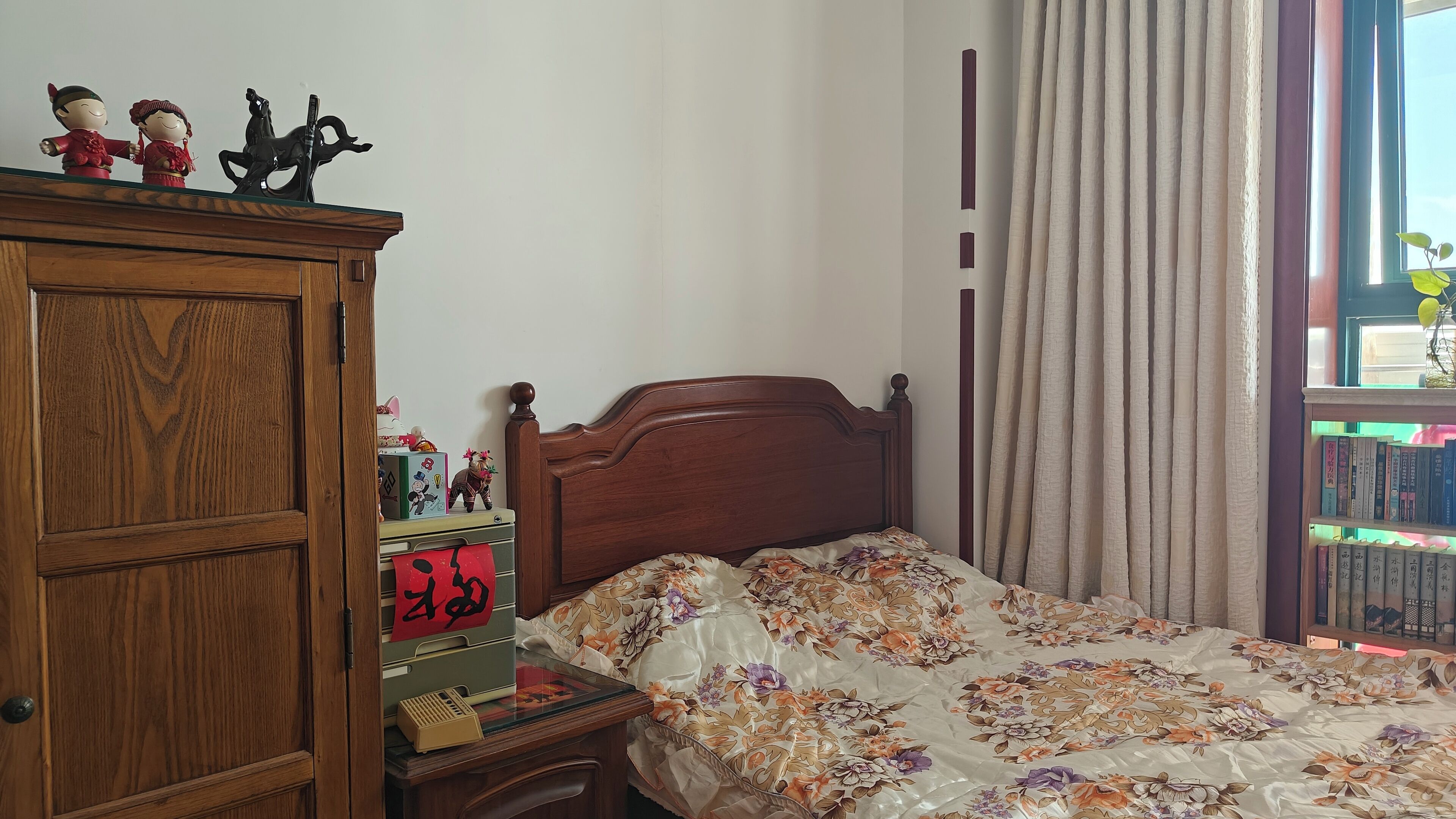 Un dormitorio con un armario de madera, una cama con ropa de cama floral, una mesita de noche con libros y una ventana con cortinas.