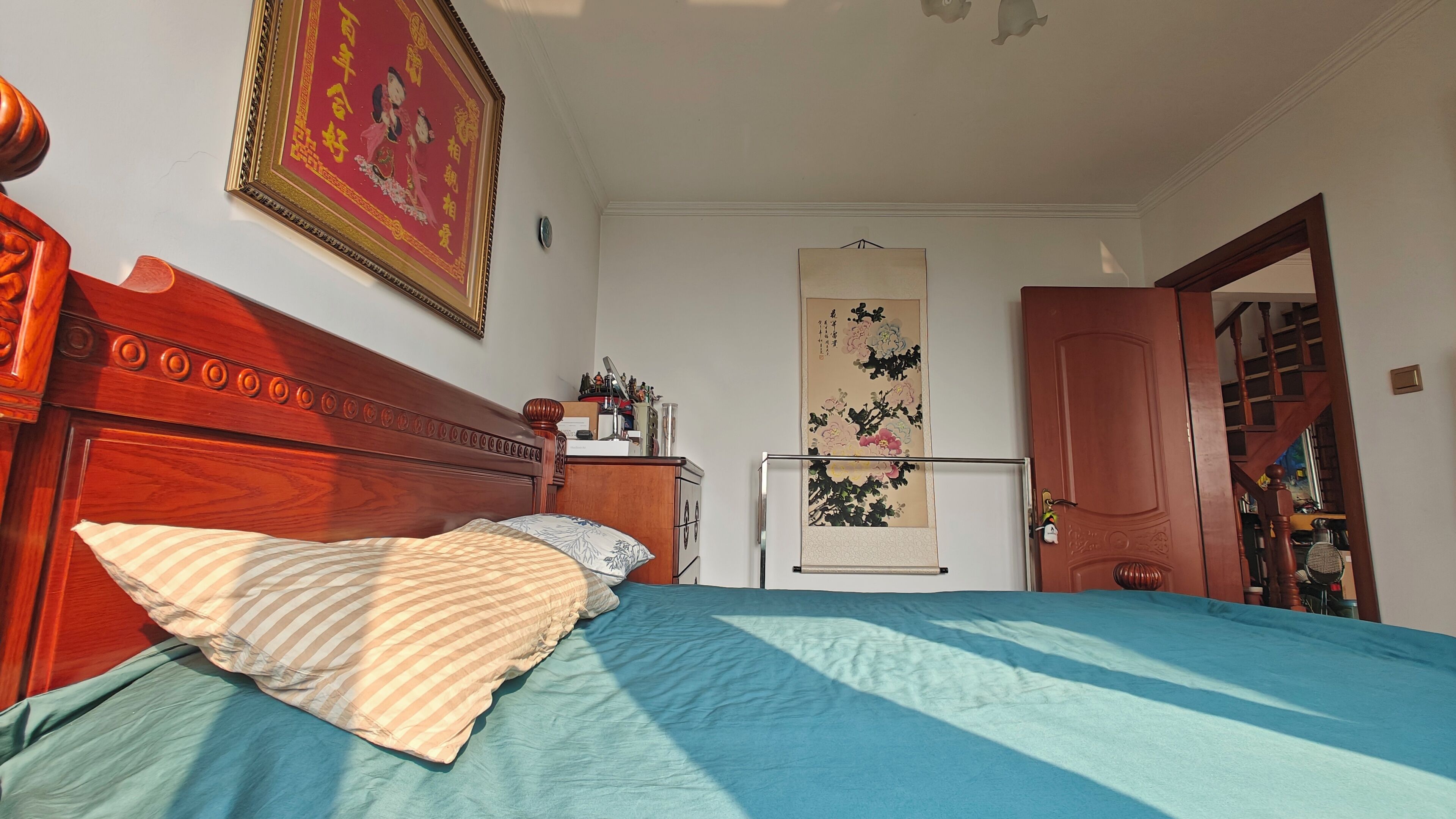 Un dormitorio con cabecera de madera, cama con ropa de cama azul, cómoda con adornos, pared con un cuadro enmarcado y una puerta que da a otra habitación.