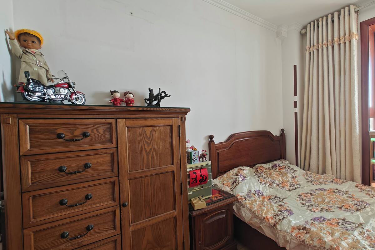 A bedroom with a wooden dresser, a bed with a floral bedspread, a nightstand with a lamp, and a window with curtains.