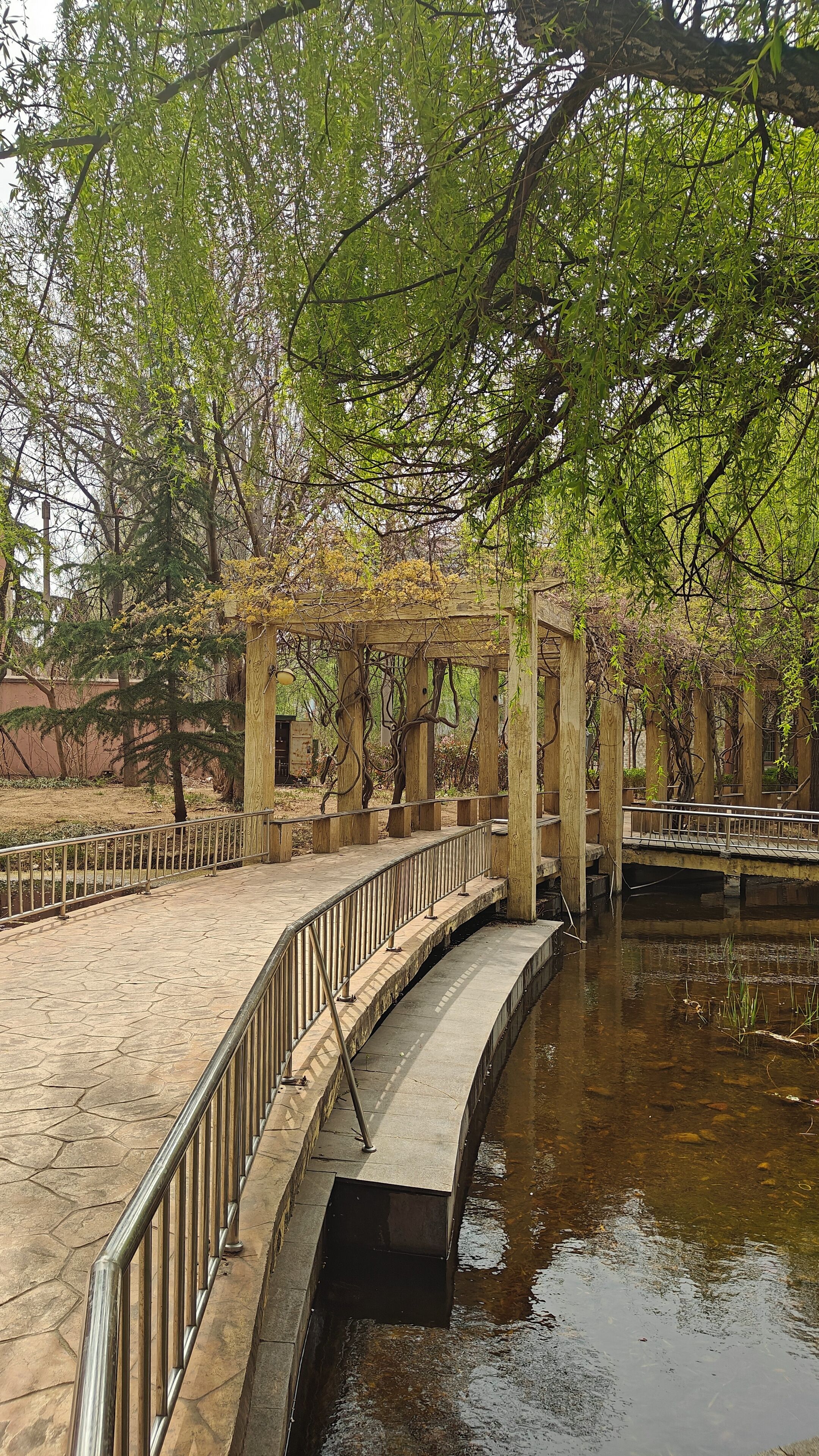 A stone bridge over a calm water body, surrounded by trees and a wooden pergola structure.