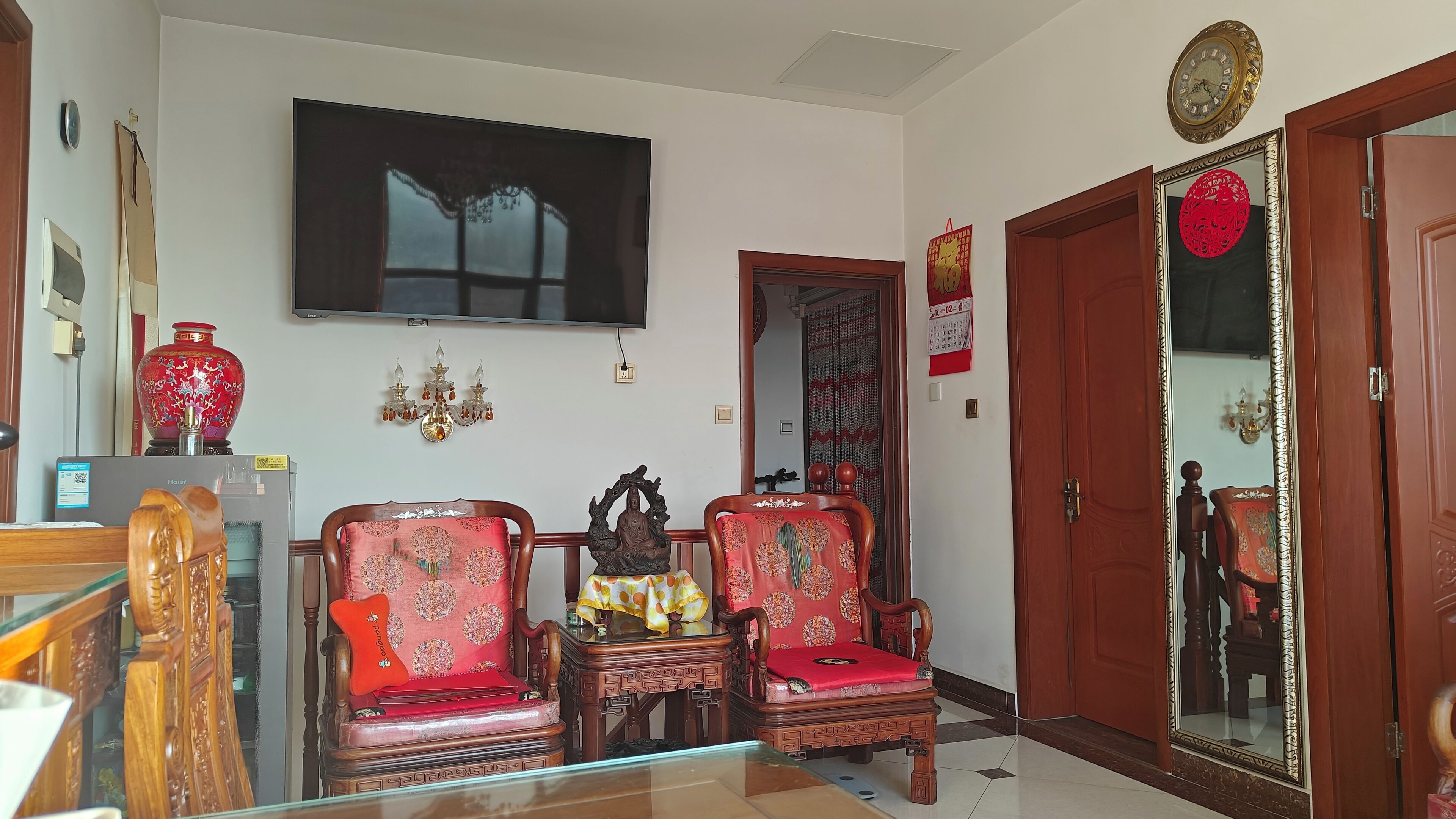 A traditional room with two ornate wooden chairs, a television, a large red vase, and a wooden door with a mirror.