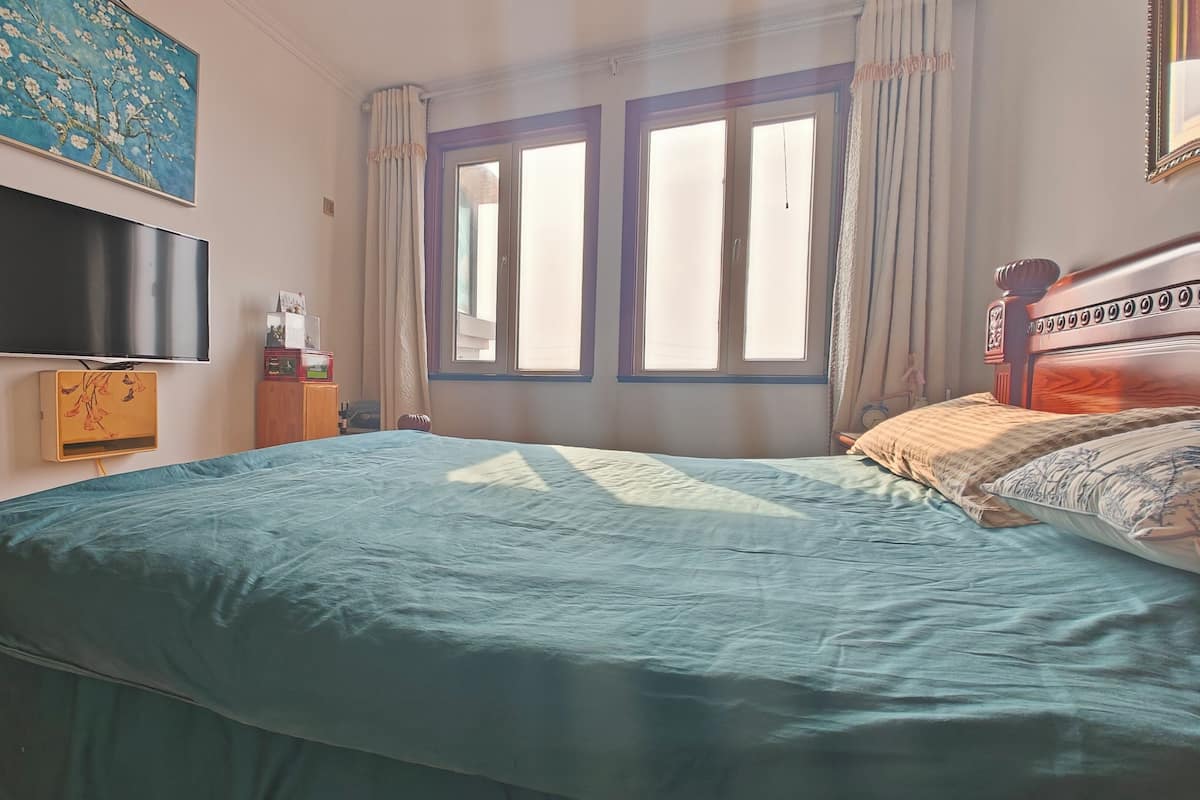 A bedroom with a large bed, a television mounted on the wall, a window with curtains, and a wooden headboard.