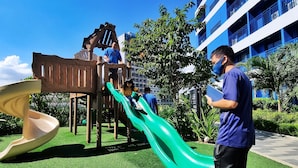 Ein Spielplatz mit einer Holzrutsche und einer grünen Rutsche, umgeben von Kunstgrün und Bäumen.