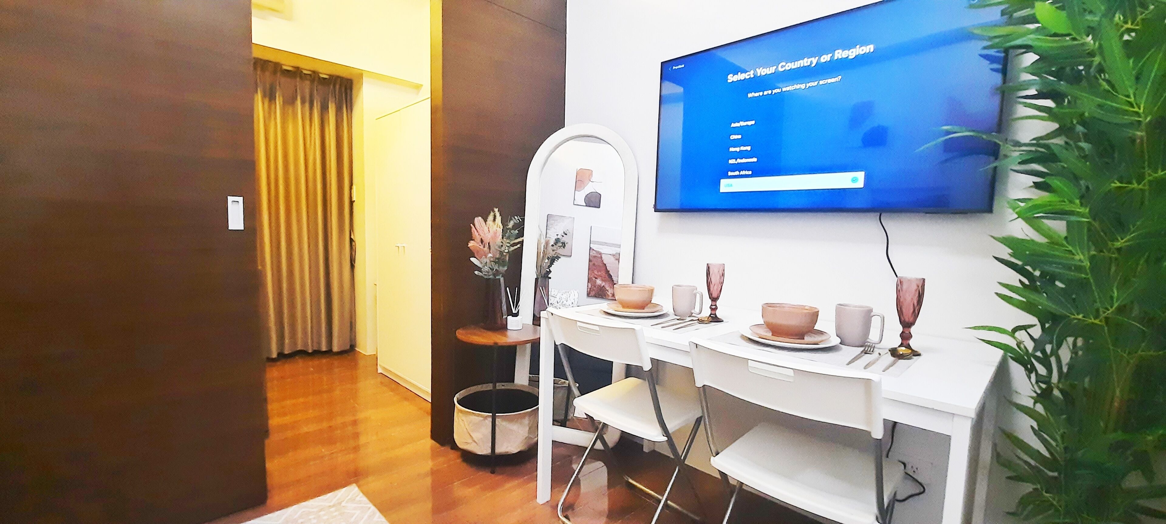 A hotel room with a dining area featuring a white table set with plates, cups, and a vase. A television is mounted on the wall displaying a menu.
