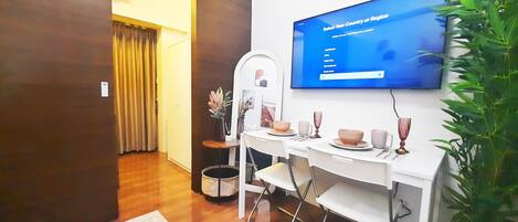A hotel room with a dining area featuring a white table set with plates, cups, and a vase. A television is mounted on the wall displaying a menu.