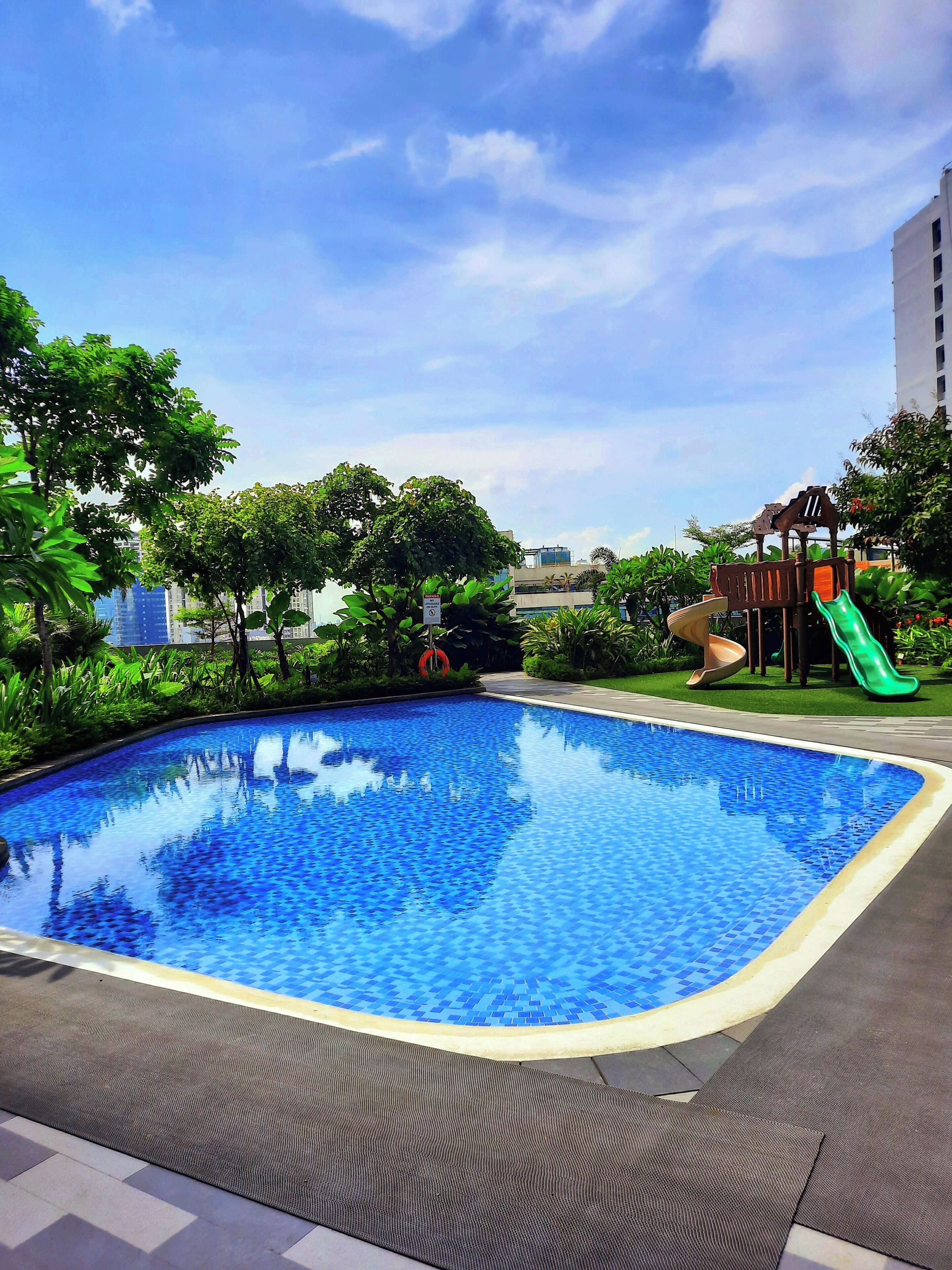 A swimming pool with a slide, surrounded by greenery and a playground.
