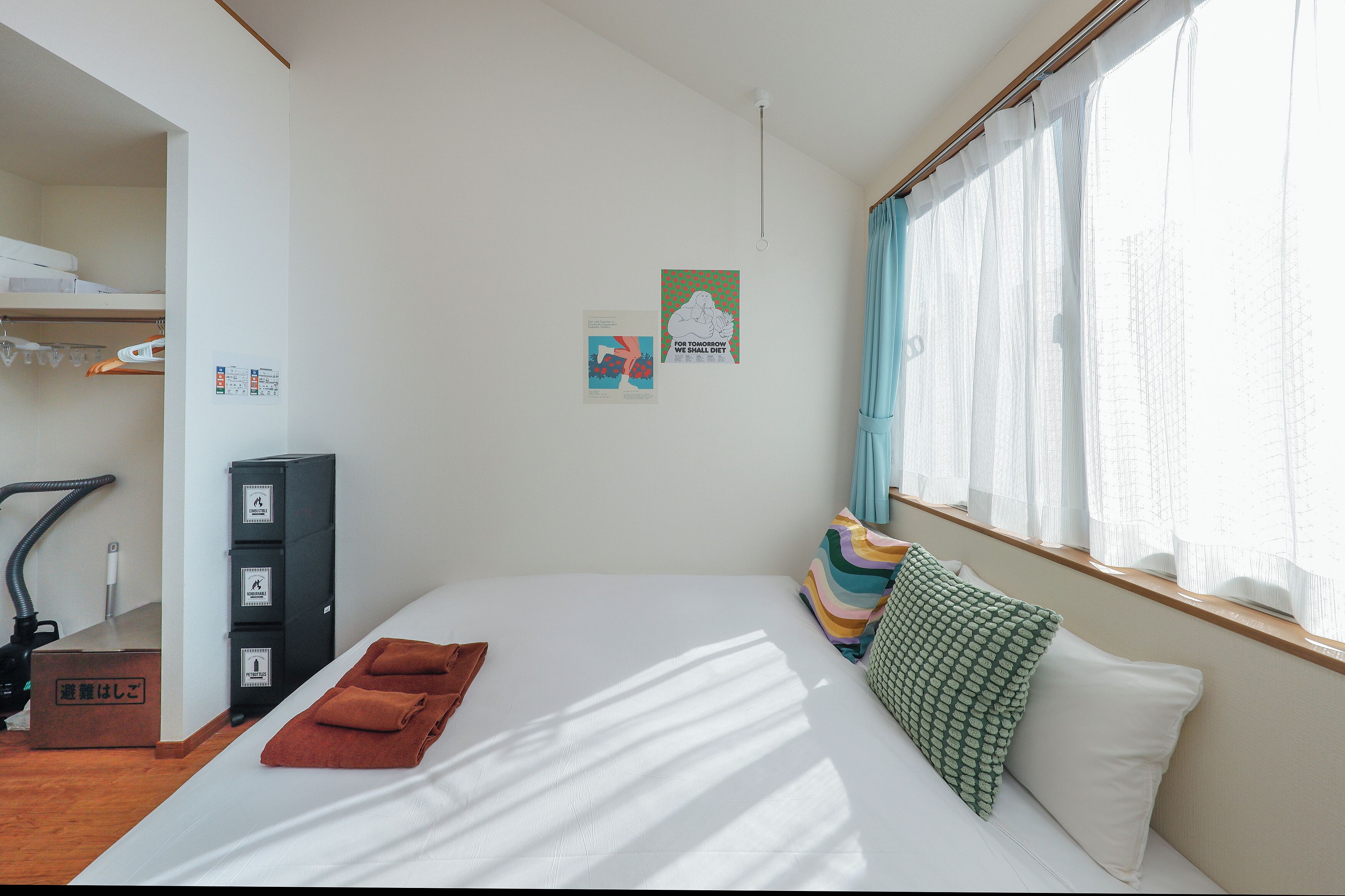 A hotel room with a bed, a window with curtains, a wall with two posters, and a closet with hanging clothes.
