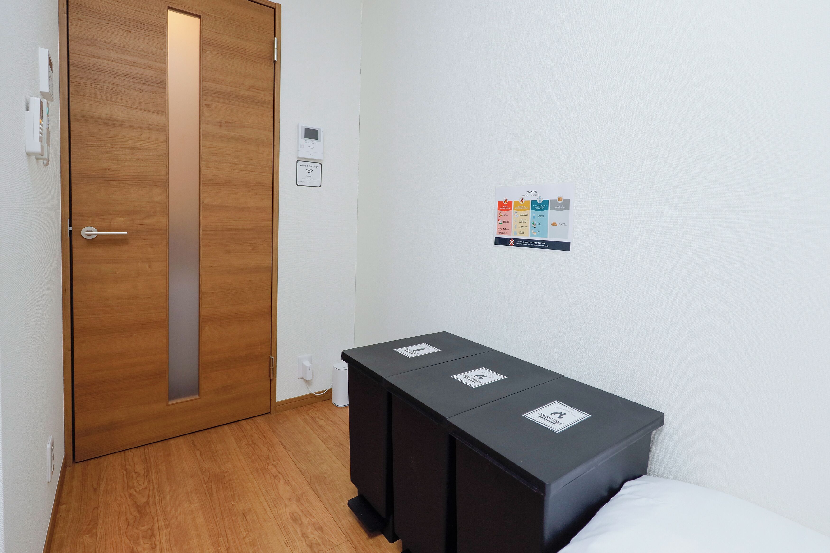 A hotel room with a wooden door, a bedside table with a black storage unit, a white wall with a Wi-Fi sign, and a small information board.