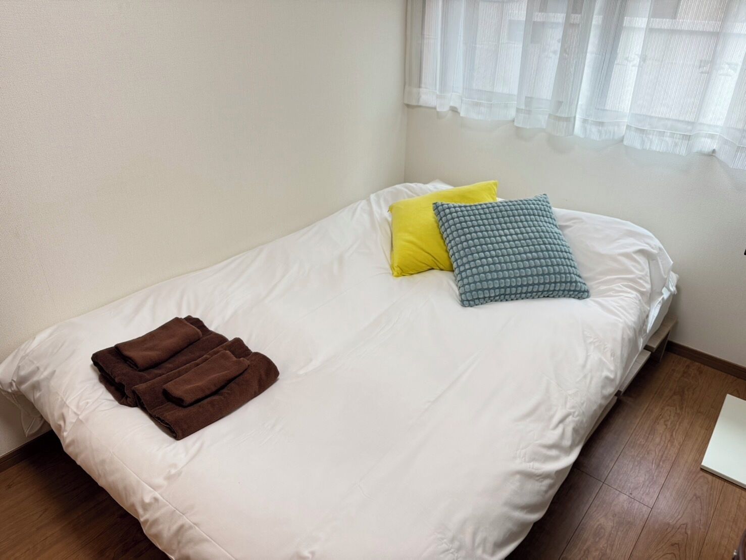 A single bed with white bedding, two pillows (one yellow, one blue), and a folded brown towel.