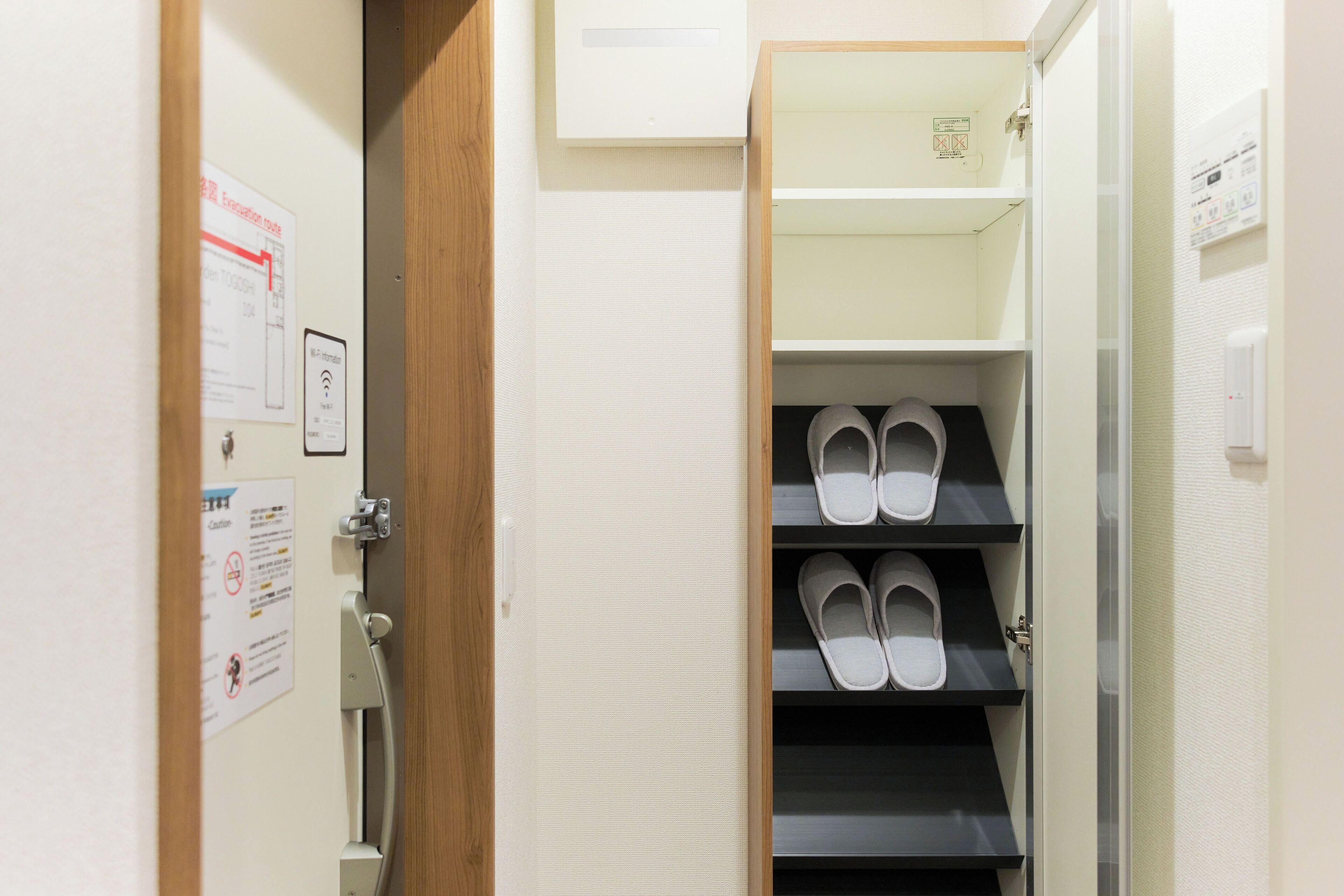 A hotel room with a wooden door, a coat rack, and a shoe rack with white slippers.