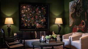 Ein Zimmer mit einer dunkelgrünen Wand, einem großen Blumenbild, einem weißen Sofa, einem beige farbenen Sessel, einem runden Couchtisch mit einer Kerze und zwei Tischlampen.