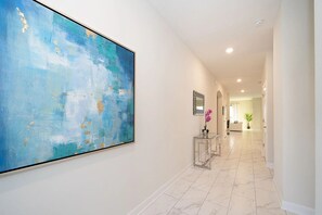 Un pasillo con un cuadro abstracto grande, una mesita de cristal con un florero y una planta en maceta.