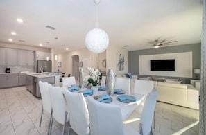 Una cocina moderna con electrodomésticos de acero inoxidable, un comedor con sillas blancas y una mesa central, y una sala de estar con un sofá y un televisor de pantalla plana.
