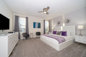 Un dormitorio moderno con una cama blanca, ventilador de techo, televisor y escritorio con cajones.