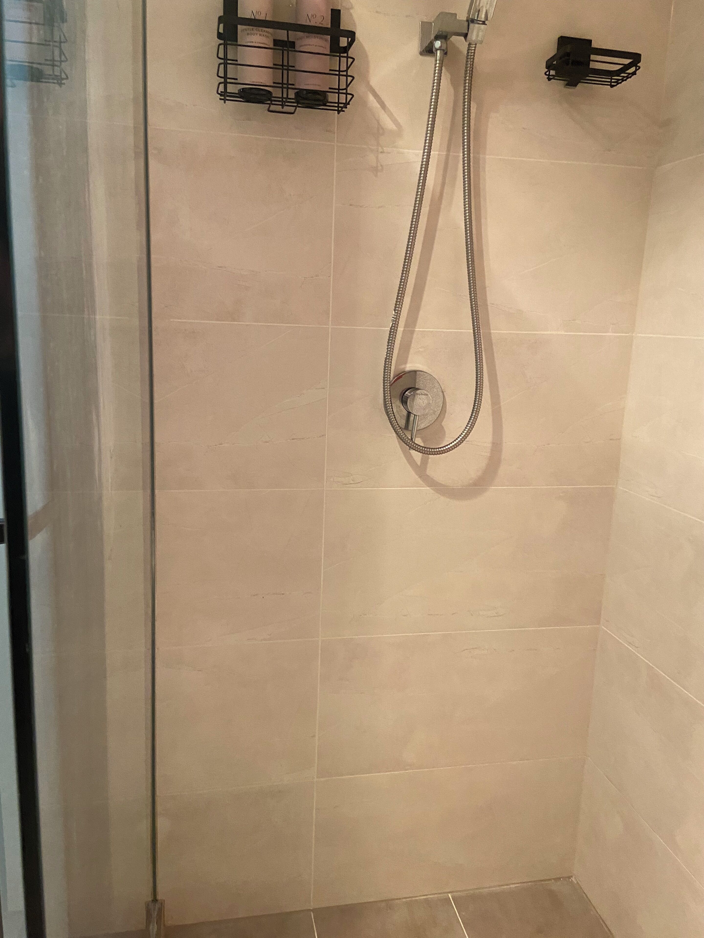 Een douche met een handdouche en een wandmengkraan. Er is een zwarte handdoekhouder met twee flessen erop.