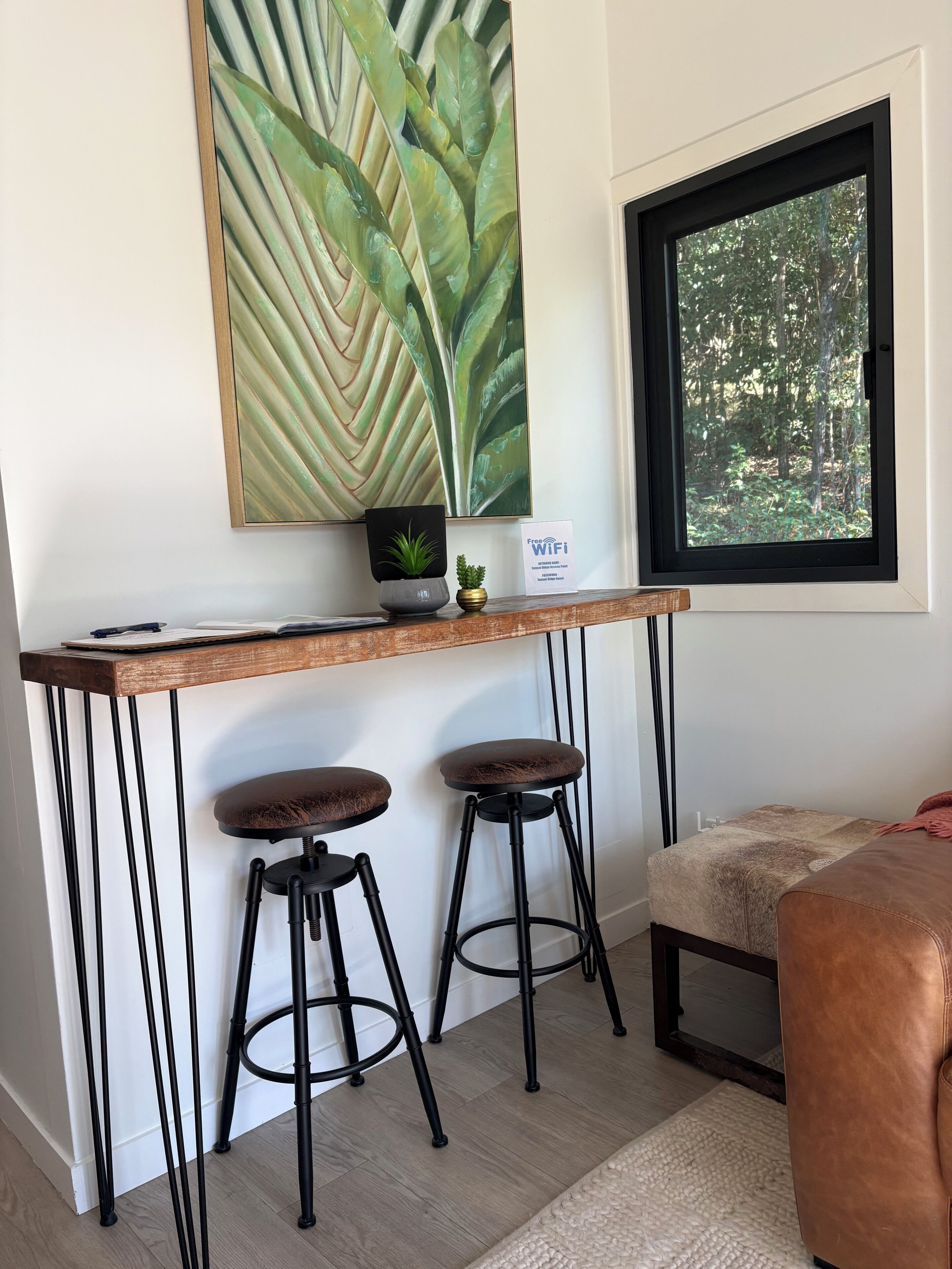 A small, modern room with a wooden desk, two stools, a large window with a view of trees, and a wall-mounted plant.