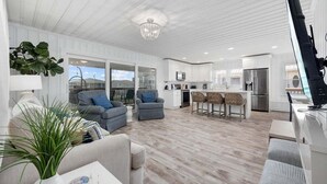 A modern living room with a large window, a dining area with a bar, and a kitchen with white cabinetry. - Summer Breeze | 5BR Waterfront Home w Heated Pool (North Myrtle Beach)