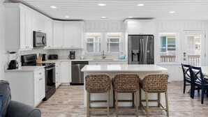A modern kitchen with white cabinetry, stainless steel appliances, and a central island with bar stools. - Summer Breeze | 5BR Waterfront Home w Heated Pool (North Myrtle Beach)