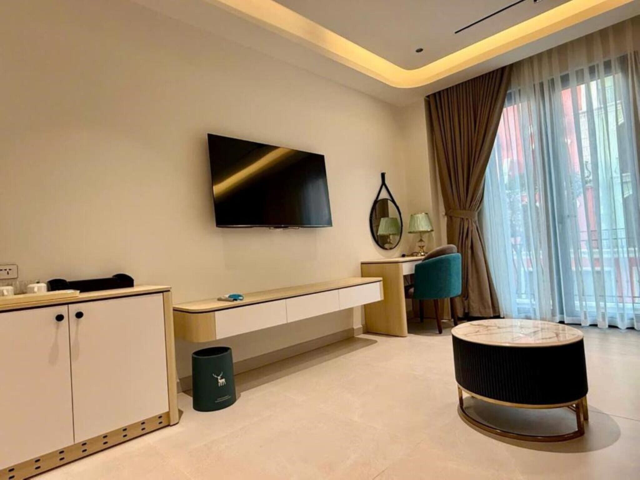 A modern hotel room with a flat-screen TV mounted on the wall, a desk with a chair, a round coffee table, and a large window with curtains.