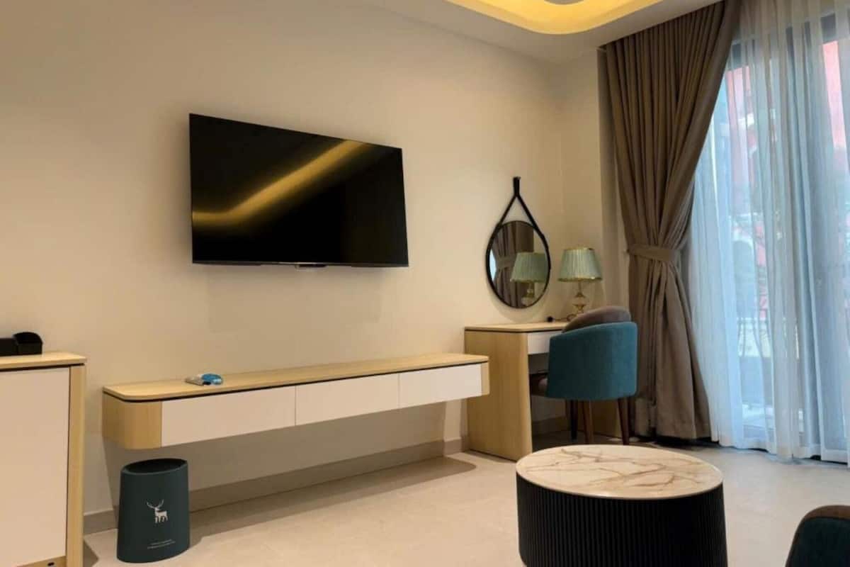 Une chambre d’hôtel moderne dotée d’un grand téléviseur à écran plat fixé au mur, d’une console en bois, d’une table basse ronde en marbre et offrant une vue sur la ville à travers de fines rideaux.