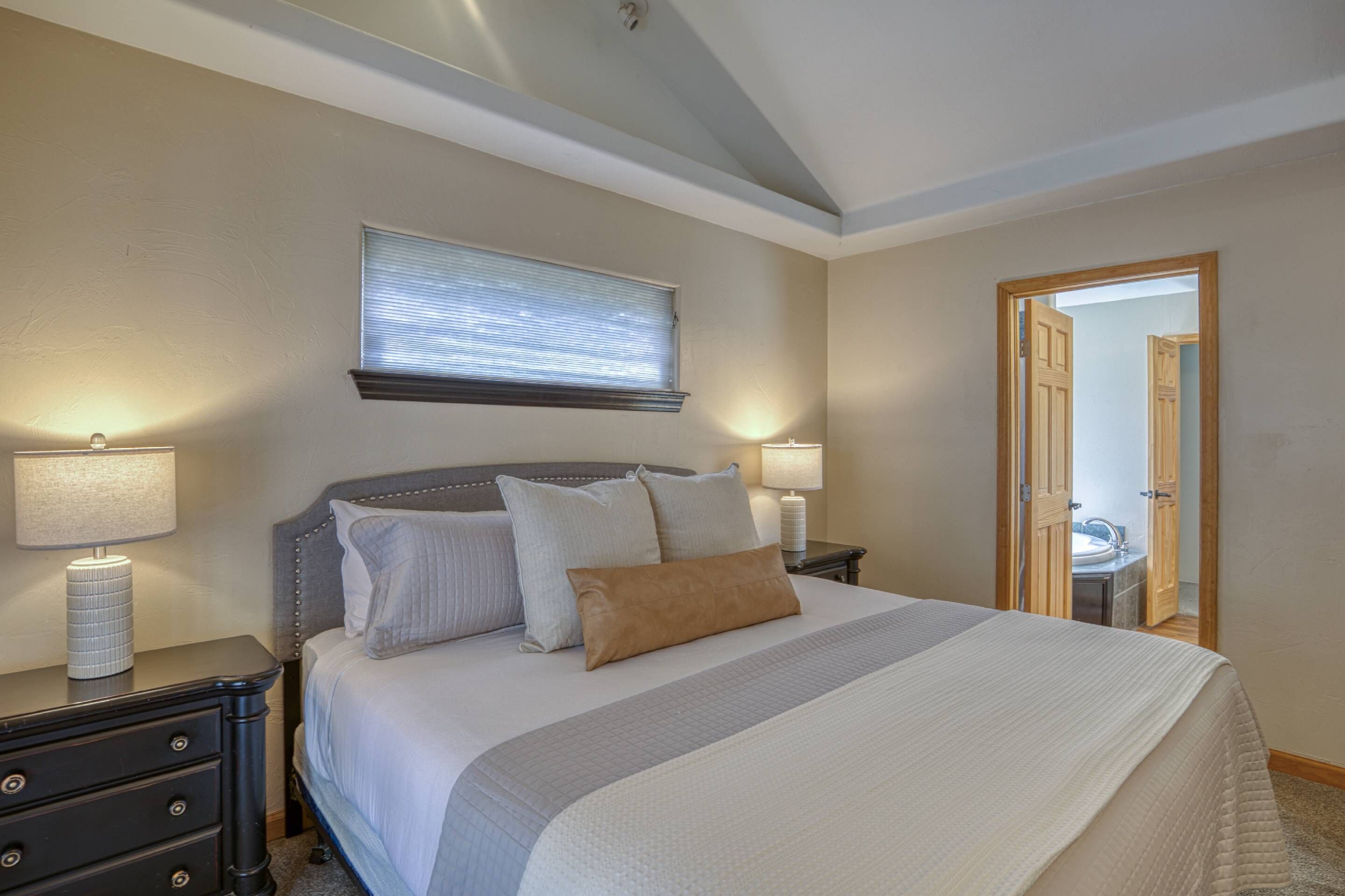 A neatly arranged bedroom with a large bed, two bedside tables with lamps, a window with blinds, and a door leading to another room.