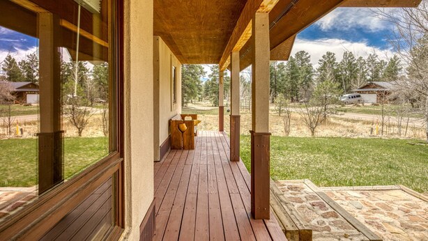 A covered porch with a wooden floor, large windows, and a view of a grassy area with trees and a distant building. - Mountain Views, Pet Friendly, New Hot Tub, Fenced Yard, Near Trails! (Pagosa Springs)