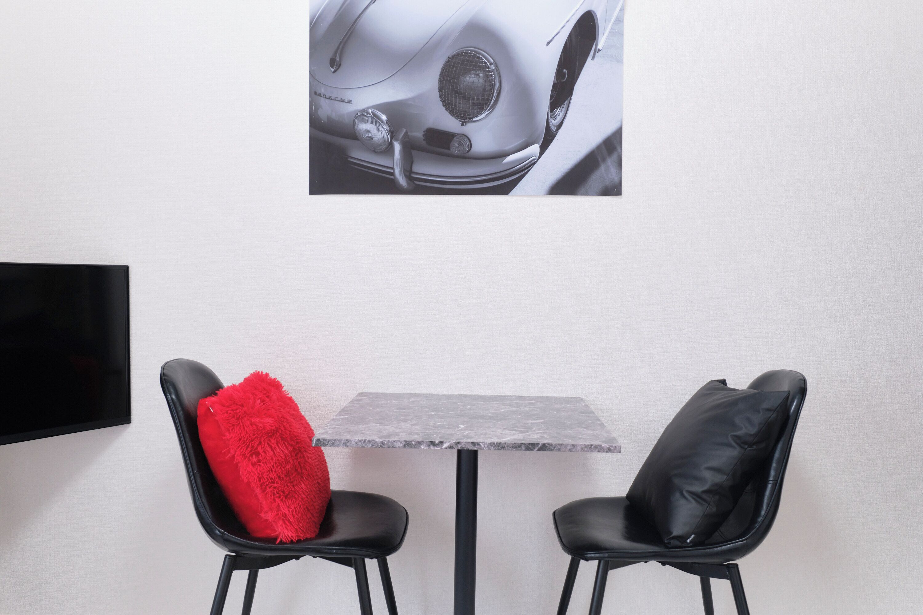 A room with a grey marble table, two black chairs, one with a red cushion, and a wall-mounted TV.