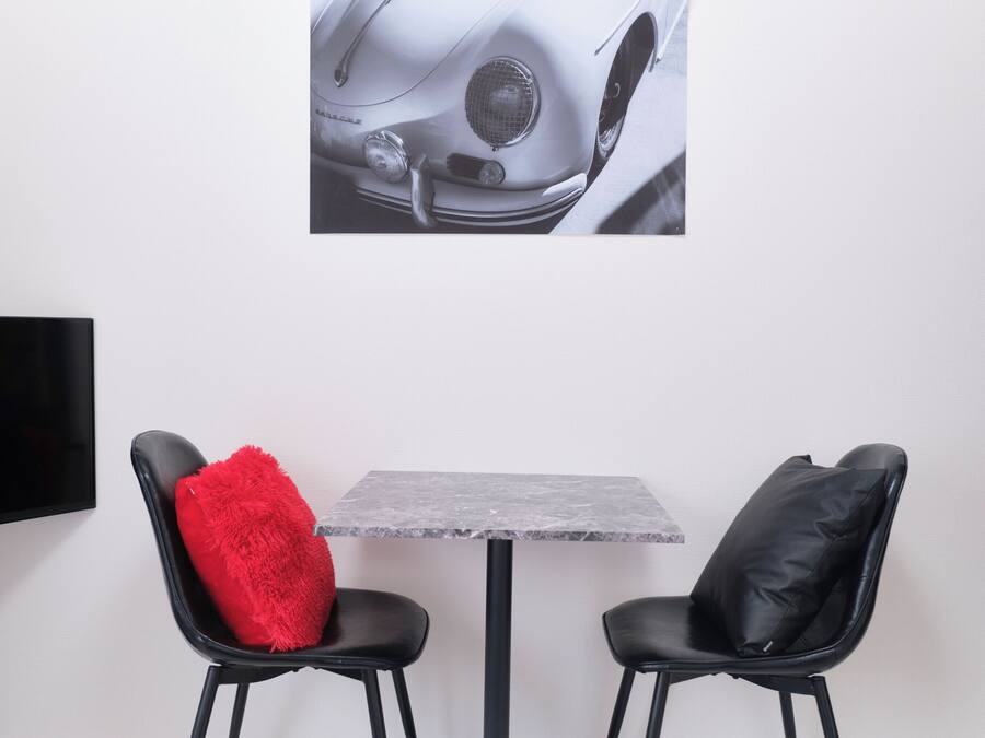 A room with a grey marble table, two black chairs, one with a red cushion, and a wall-mounted TV.