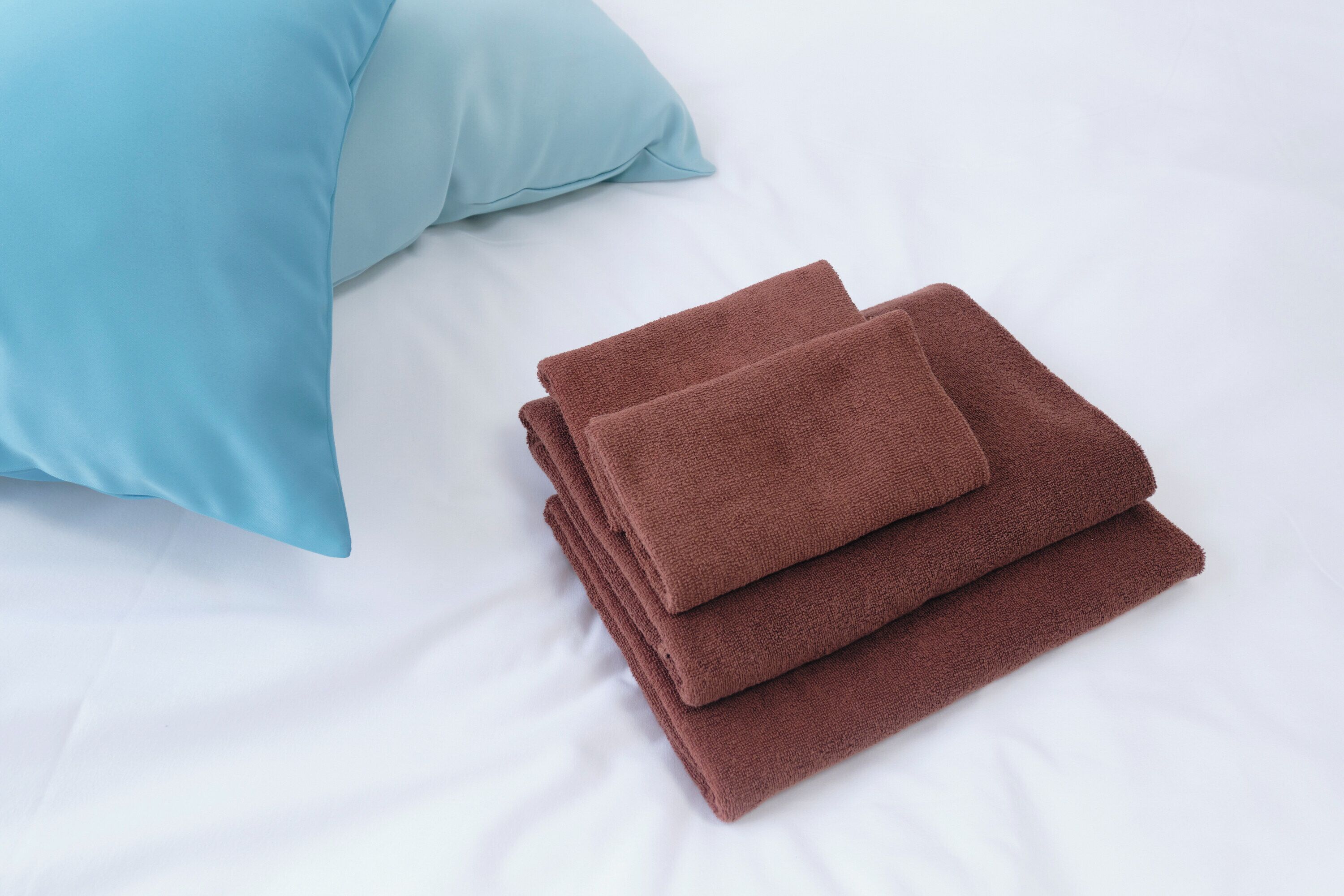 Une serviette marron soigneusement pliée et un oreiller bleu clair sur un lit blanc.