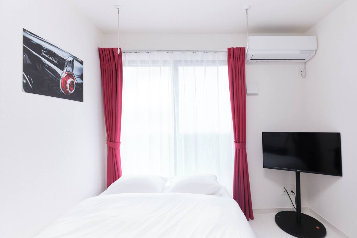 A hotel room with a bed, a flat-screen TV, an air conditioner, and a window with sheer curtains.