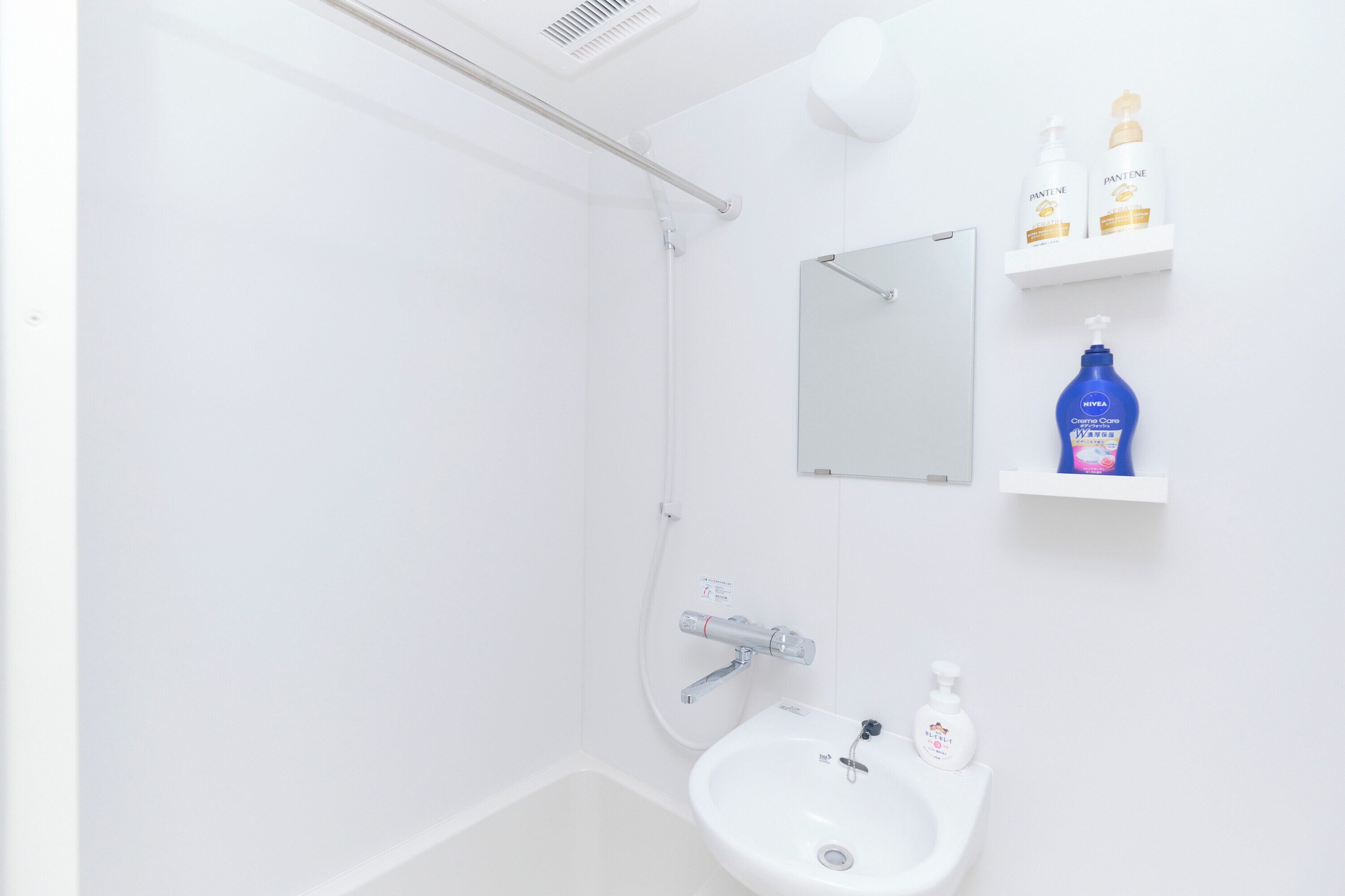 Een badkamer met een wit wastafel, een spiegel en een douchegedeelte. Toiletartikelen zijn aan de muur bevestigd en er staat een blauwe fles Nivea Creme Care.