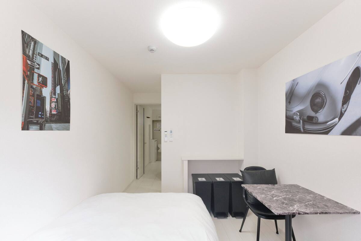 A small, clean room with a bed, a desk, and a chair. There are two trash bins and a door leading to another room.