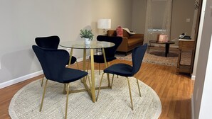 Ein Essbereich mit einem Glastisch, umgeben von vier dunklen Stühlen mit goldenen Beinen, einer kleinen Topfpflanze und einer Lampe auf einem Beistelltisch.