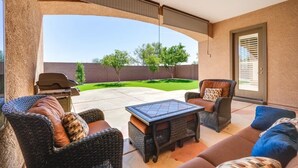 Un patio couvert aménagé avec des meubles en osier, une table à plateau en verre et une vue sur un jardin arrière paysager.
