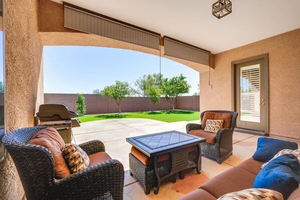 Uma varanda coberta com móveis de vime, uma mesa de vidro e vista para um jardim bem cuidado.